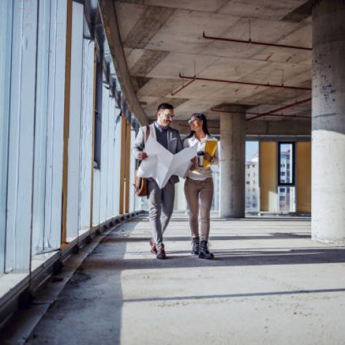 Full length of attractive caucasian architects walking in future business center and talking about fresh ideas for building. Man holding blueprints while woman holding paperwork.